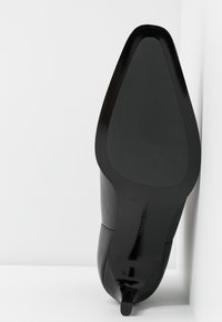Chaussure à talon haut noire reposant semelle vers le haut dans un coin blanc, montrant un bout pointu et un talon fin.