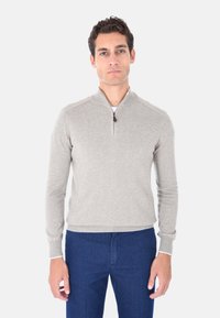 Felpa grigia chiara con zip e colletto alto, caratterizzata da cuciture a contrasto e polsini a coste, abbinata a pantaloni blu scuro.