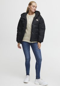Svart puffjakke med hette, med glidelås foran og lommepaneler. Bært over en kremfarget strikkegenser og blå jeans med hvite sneakers.