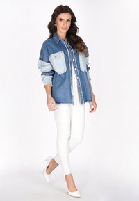 Denim oversized shirt met lichte en donkere blauwe secties, twee borstzakken en een gerafelde onderkant, gecombineerd met witte skinny pants en hakken.