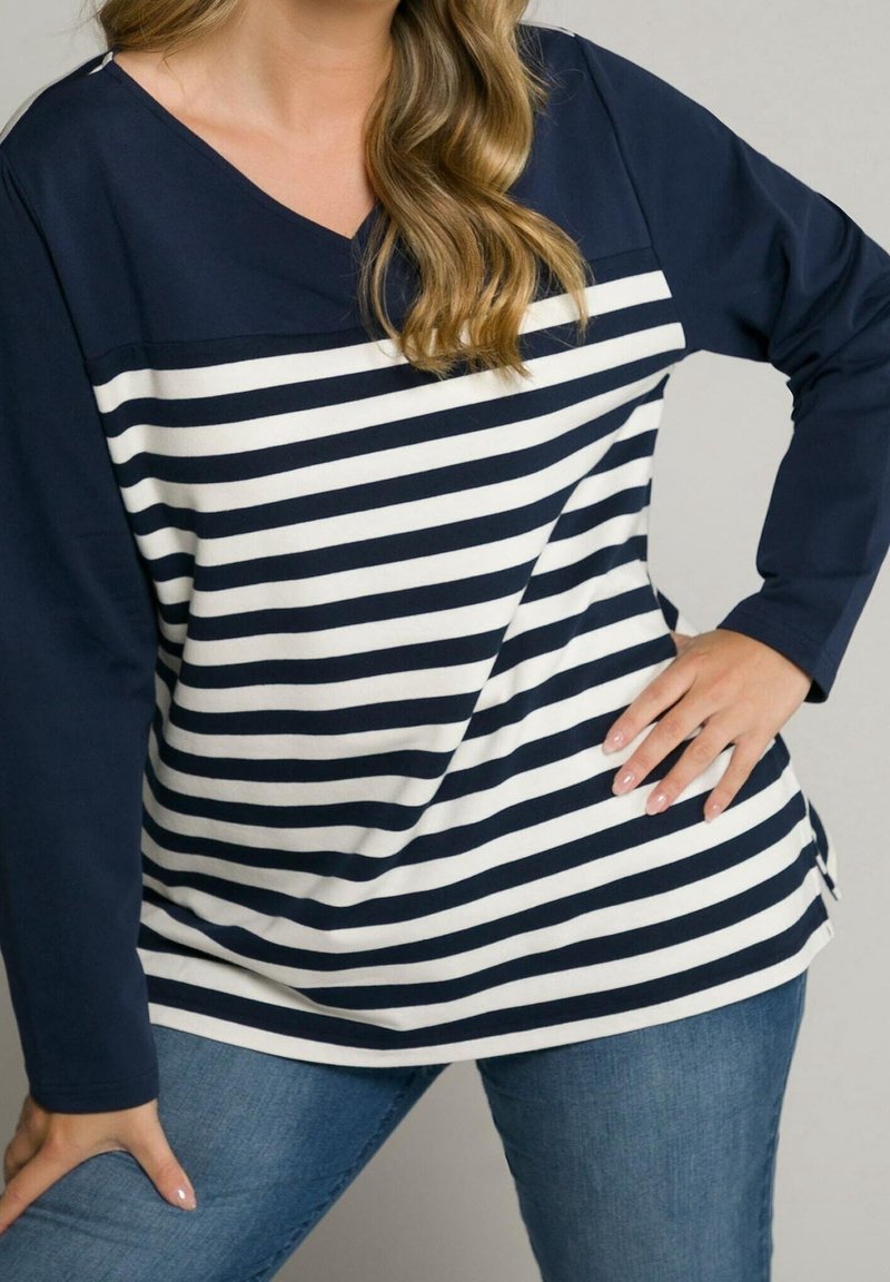 Donna che indossa una maglietta a righe orizzontali blu navy e bianca con maniche blu navy e jeans blu, in posa con le mani sui fianchi e sulla coscia.