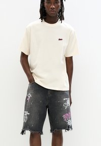 T-shirt crema con collo a giro e un piccolo logo rosso, abbinata a pantaloni bermuda in denim nero strappati con grafiche colorate e orli sfilacciati.