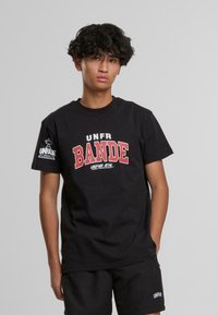 Juodas medvilninis marškinėliai su ryškiu raudonu ir baltu „UNFR BANDE“ dizainu priekyje ir logotipu kairiajame rankove. Trumpi rankoviai, apvalus kaklas.