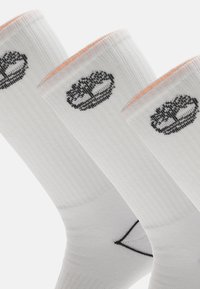 Weiße Socken mit gerippter Textur, rosa Akzenten an der Oberseite und einem schwarzen Logo mit einem Baumdesign. Zwei Paare gezeigt.