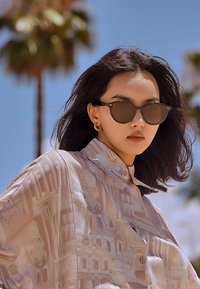 Una camicia beige chiaro con motivi architettonici, abbinata a occhiali da sole scuri, sullo sfondo di un cielo azzurro e palme.