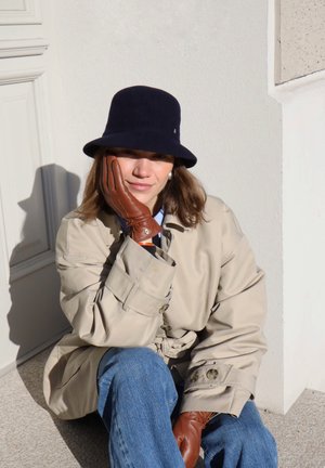 Beige trench coat with rolled sleeves, blue shirt underneath, blue felt bucket hat, and brown leather gloves, sitting against a white wall.