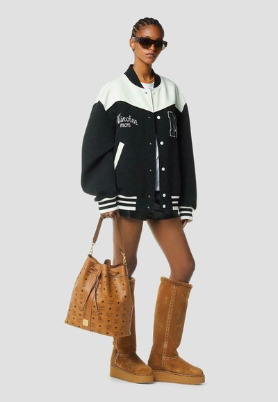 Femme portant une veste varsity noire et blanche surdimensionnée, des bottes hauteur genou en daim marron, des lunettes de soleil et tenant un sac à main en cuir marron à motif.