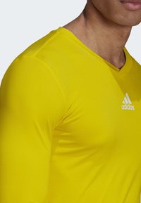 Man som bär en åtsittande gul Adidas långärmad tröja med synlig varumärkeslogotyp på bröstet mot en neutral bakgrund.