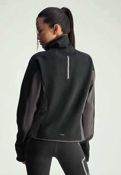 Giacca sportiva nera e grigia con colletto alto, tessuto testurizzato e dettagli riflettenti. Presenta un design elegante e una silhouette aderente.