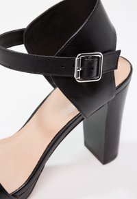 Sandale noire à talon haut avec talon épais, bride de cheville et boucle argentée sur semelle intérieure beige pâle sur fond blanc.