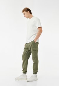 Weißes T-Shirt mit kurzen Ärmeln, kombiniert mit olivgrünen Cargo-Hosen mit Seitentaschen und elastischen Bündchen, ergänzt durch weiße Sneakers.