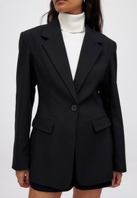 Juodas blazeris su pritaikytu siluetu, tekstūruotu audiniu, iškirptomis atlapomis, viena sagtele ir dviem priekinėmis kišenėmis, derinamas su baltu simpatišku megztiniu.