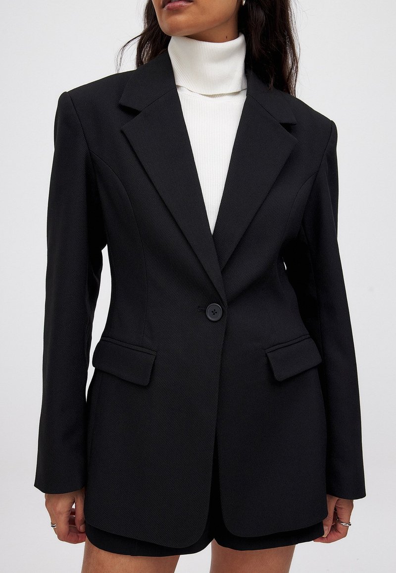 Juodas blazeris su pritaikytu siluetu, tekstūruotu audiniu, iškirptomis atlapomis, viena sagtele ir dviem priekinėmis kišenėmis, derinamas su baltu simpatišku megztiniu.