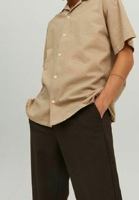 Persona che indossa una camicia beige a maniche corte con texture, sbottonata e lasciata all'esterno, sopra dei pantaloni neri, con le mani nelle tasche davanti a uno sfondo neutro.