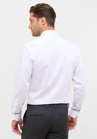 Witte blouse met lange mouwen en een standaard kraag. Heeft mouwen met knopen en contrasterende biezen. Draagt bij grijze broek, wat zorgt voor een getailleerde pasvorm.