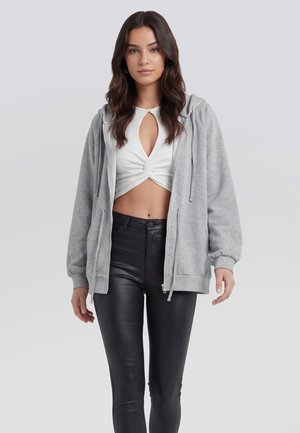 Frau mit langen dunklen Haaren, die einen hellgrauen Zip-Up-Hoodie, ein weißes, bauchfreies Top mit Knoten vorne und schwarze, hoch taillierte Lederhosen trägt.