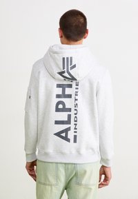 Svetlošedá mikina s kapucňou s veľkým čiernym logom "ALPHA INDUSTRIES" na chrbte, s vačkovým vreckom a pružnými manžetami.