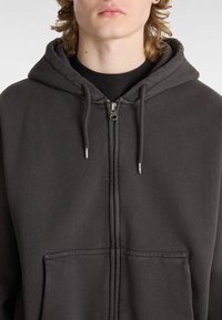 Grauer Reißverschluss-Hoodie aus weichem Stoff, mit einer großen Fronttasche, Kordeln und einer gefütterten Kapuze. Minimalistisches Design mit sichtbaren Nähten.