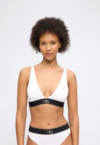 Γυναίκα με κοντά σγουρά μαλλιά, φορώντας λευκό σουτιέν και αντίστοιχο βρακάκι Calvin Klein με μαύρες ελαστικές ταινίες με το σήμα της εταιρείας, στέκεται μπροστά σε απλό φόντο.