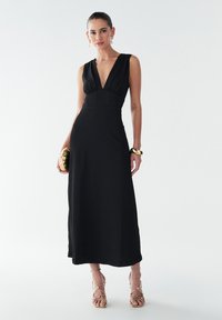 Robe maxi noire sans manches avec un décolleté en V profond, tissu texturé, corsage ajusté et jupe évasée. Accessoirisée avec des bracelets en or et un sac à main.