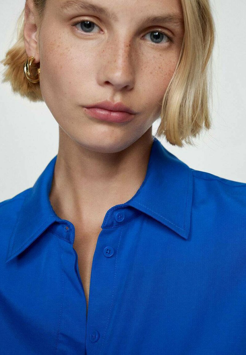 Chemise bleu à col avec un devants boutonné, présentant une texture lisse. Le modèle porte de grandes boucles d'oreilles en or et a les cheveux blonds courts.