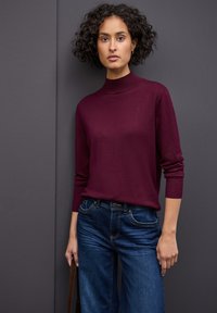 Pull en col roulé bordeaux avec des poignets côtelés, associé à un jean bleu foncé doté de poches avant et d'une coupe ajustée.