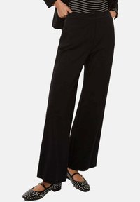 Pantaloni neri a gamba larga con una texture liscia, dotati di vita alta e tasche frontali. Abbinati a ballerine nere con borchie.