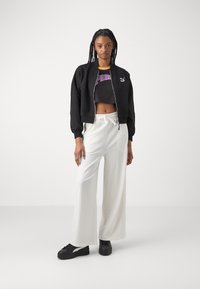 Veste noire courte, top court gris avec un graphique "Puma", pantalon large blanc, et baskets noires avec des accents de rayures blanches.