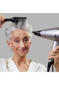 Ασημί πιστολάκι μαλλιών με κομψό σχέδιο και μαύρη μύτη. Το μοντέλο κρατάει μια μαύρη στρογγυλή βούρτσα ενώ περιποιείται τα μαλλιά. Ο γκρι φόντος ενισχύει την αντίθεση.