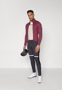 Radbekleidung mit einer weinrot leichten Jacke, einem beigen Oberteil, schwarzen gepolsterten Hosen mit weißen Akzenten und weißen Radschuhen. Hält einen schwarzen Helm.
