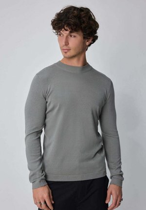 Jeune homme aux cheveux bouclés et foncés, portant un pull gris moulant à manches longues et un pantalon noir, regardant légèrement sur le côté.