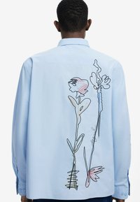 Light blue long-sleeve shirt featuring colorful floral embroidery on the back, with outlined flowers in pink, green, and blue thread.