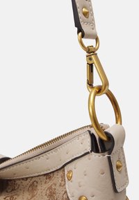 Primo piano dell'angolo di una borsa beige texturizzata con cerniera dorata, rivetti e un gancio metallico che collega la tracolla all'anello della borsa.
