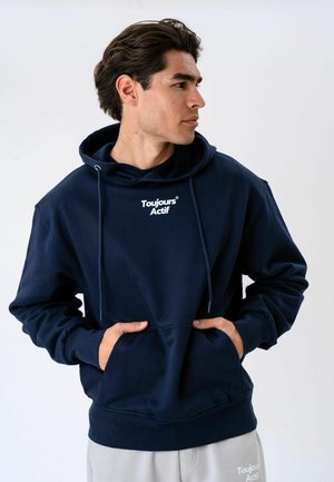 Mężczyzna ubrany w granatową bluzę z kapturem z napisem „Toujours Actif” na piersi oraz jasnoszare spodnie, patrzy w bok, trzymając ręce w kieszeniach bluzy.