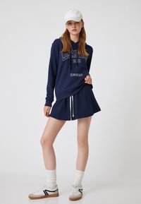 Marinblå sweatshirt med tryck "Los Angeles 1981", i kombination med en matchande kjol. Vit keps, textilmed tyg, dragsko detaljer och sneakers.