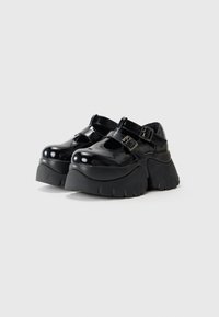 Sapatos de plataforma em verniz preto com solas de borracha largas, apresentando duas alças ajustáveis e um design de bico redondo.