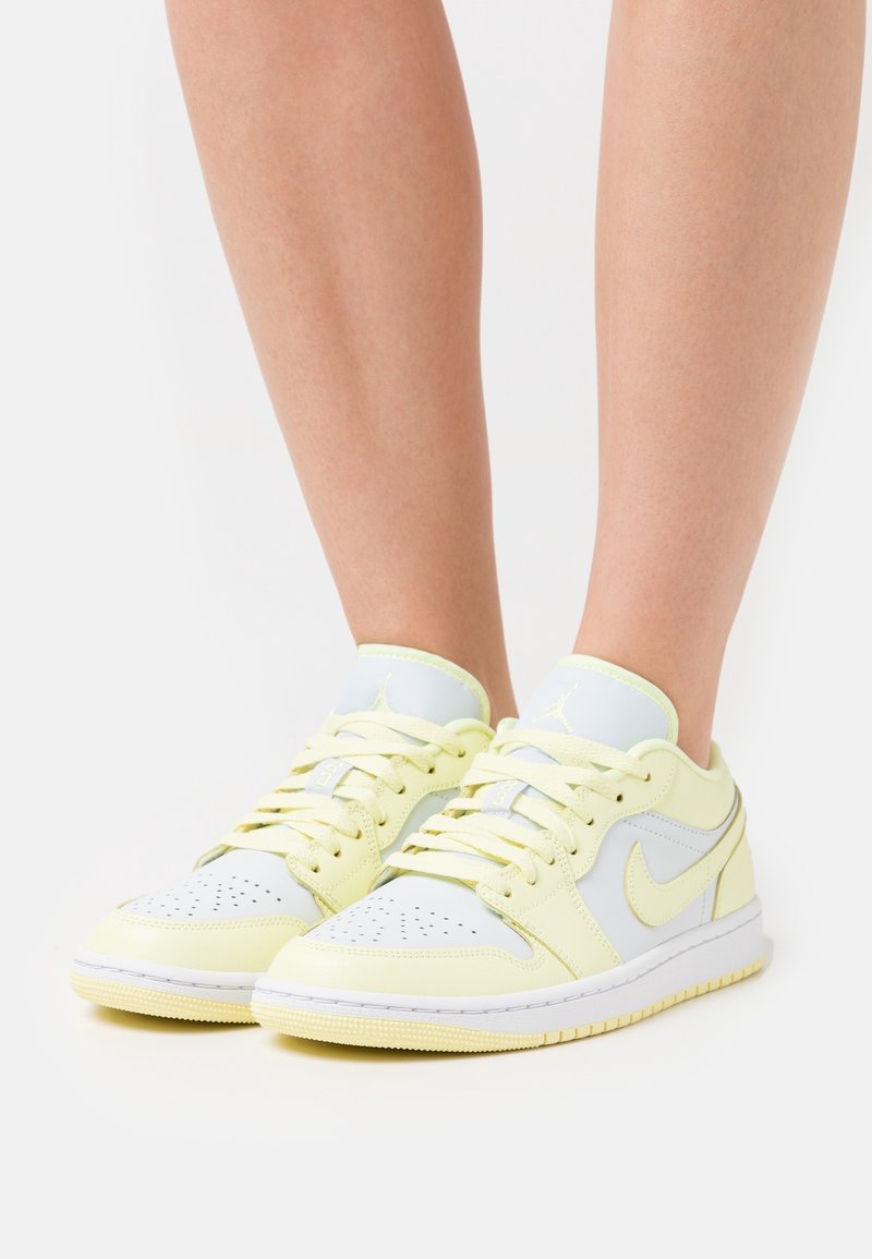 Personne portant des baskets Nike Air Jordan jaune clair et blanc avec des lacets, debout sur une surface blanche.