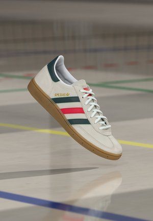 Bijela Adidas Spezial tenisica s zelenim i crvenim prugama, gumeni potplat i bijele vezice, prikazana kako lebdi iznad poda zatvorenog sportskog terena.
