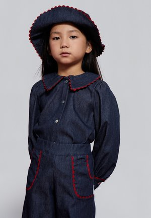 Conjunto de mezclilla que incluye una camisa con cuello y pantalones de cintura alta. Detalles en rojo con bordes ondulados en el cuello y los bolsillos; sombrero de ala ancha a juego.