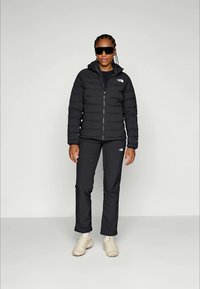 Kvinna i svart North Face-puffjacka och byxor, vita sneakers och stora svarta solglasögon, som står framför en neutral ljus bakgrund.