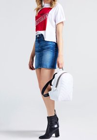 Žena v bílém a červeném zkráceném tričku, modré džínové sukni a černých kotníkových botách, držící malý bíl�ý batoh za jeho madlo.