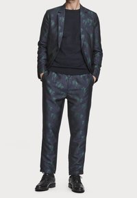 Mężczyzna w ciemnym garniturze z zielonym nadrukiem palm, czarnym swetrze pod spodem, czarnych eleganckich butach, stojący z rękami w kieszeniach.