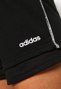Pantalones cortos deportivos negros con logo de adidas en blanco y detalles de marca, con malla en el dobladillo cerca del muslo de una persona.