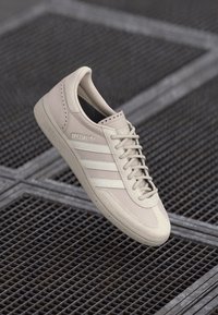 Šviesiai kreminės spalvos sportiniai bateliai su lygiu zomšiniu paviršiumi, balta trimis juostelėmis dekoracija, tekstūruotu padu ir minimalistiniu raištelių užsegimu.