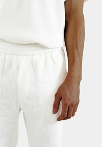 Weiße Leinenhose mit elastischem Bund, die eine leicht strukturierte Oberfläche aufweist. Eine Hand liegt lässig an der Seitennaht.