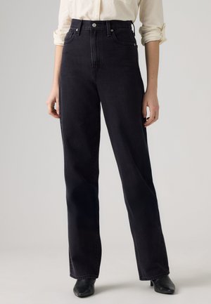 Femme portant un jean noir taille haute, coupe droite, avec bouton et fermeture éclair, associé à des chaussures noires à talons et une chemise de couleur claire.