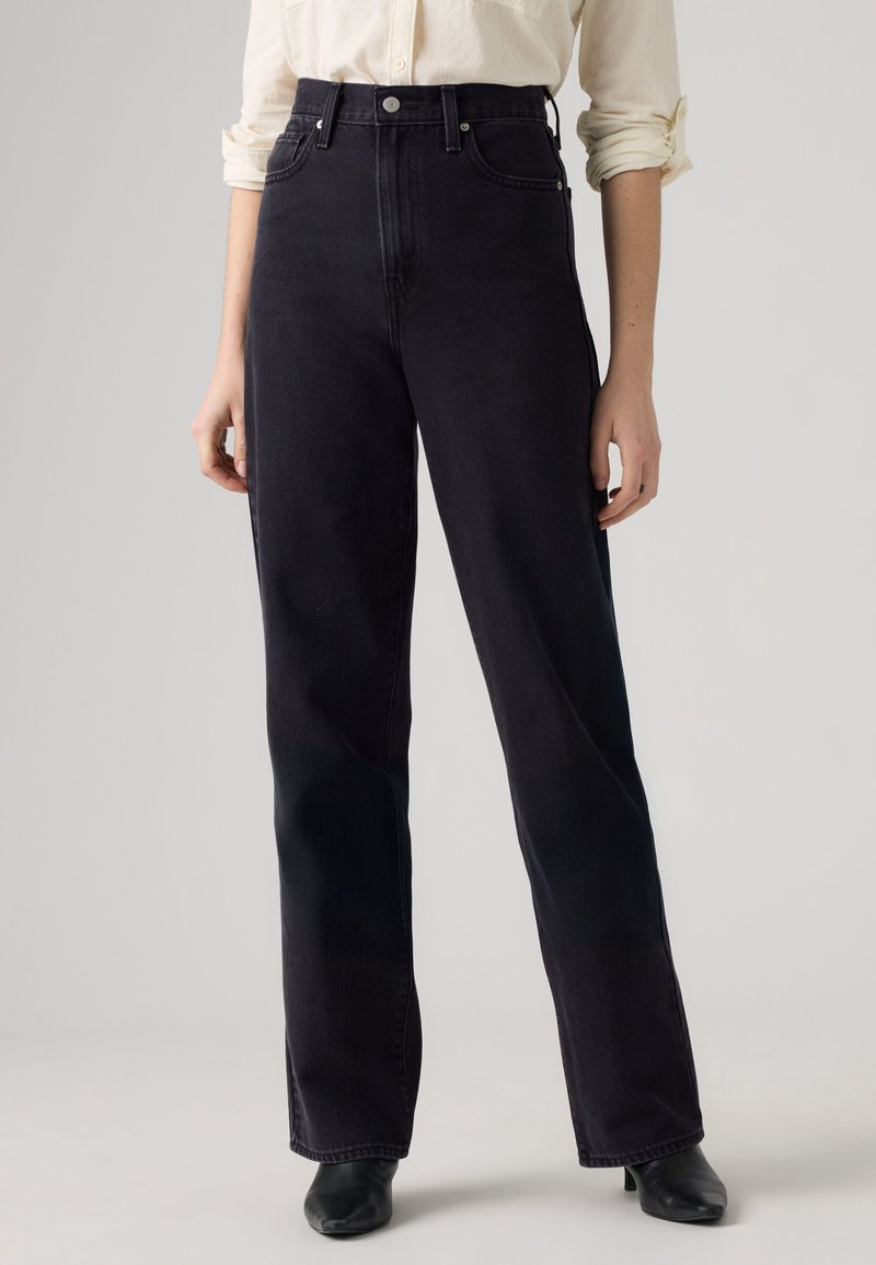 Femme portant un jean noir taille haute, coupe droite, avec bouton et fermeture éclair, associé à des chaussures noires à talons et une chemise de couleur claire.