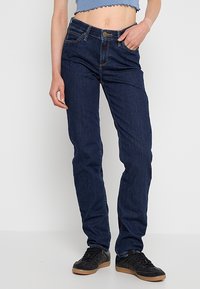 Personne portant un jean droit bleu foncé et des baskets noires, debout contre un fond clair uni, une main dans la poche.