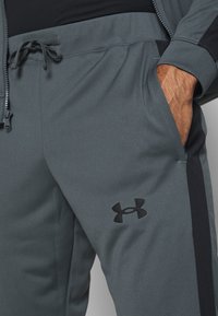 Homme portant un pantalon de survêtement gris Under Armour avec une main dans la poche et une bande latérale noire sur le pantalon.