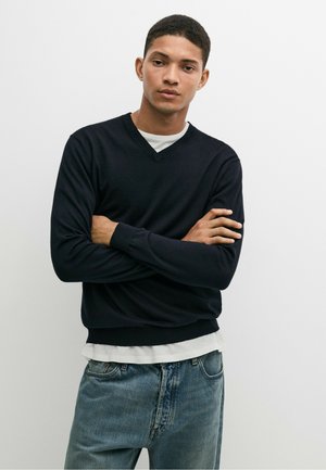 Jeune homme aux cheveux courts portant un pull noir à col en V par-dessus une chemise blanche et un jean bleu, debout les bras croisés devant un fond uni.